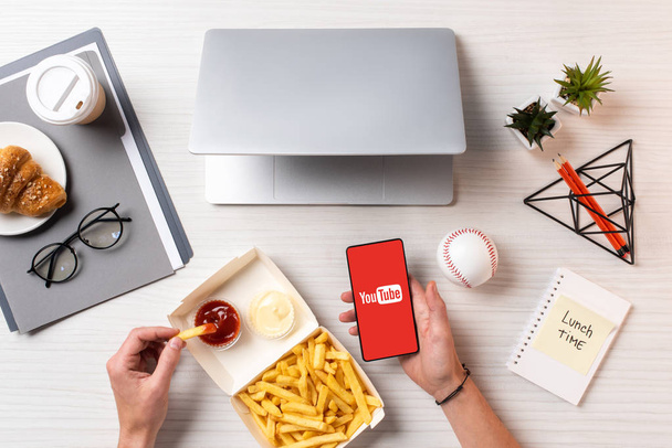 cropped shot of person eating french fries with ketchup and using smartphone with youtube app at workplace - Zdjęcie, obraz