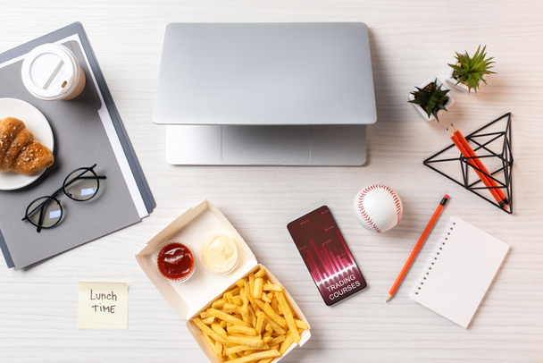 вид сверху на картофель фри, липкая записка с надписью обеденное время, ноутбук и смартфон с торговыми курсами графики на рабочем месте
 - Фото, изображение