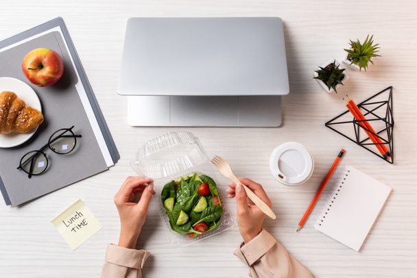 colpo ritagliato di persona che mangia insalata di verdure sul posto di lavoro con laptop e forniture per ufficio
 - Foto, immagini