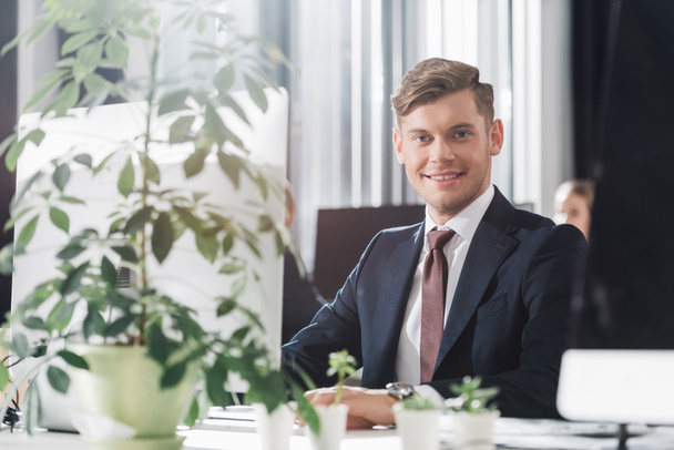 handsome young businessman sitting at workplace and smiling at camera  - Photo, Image