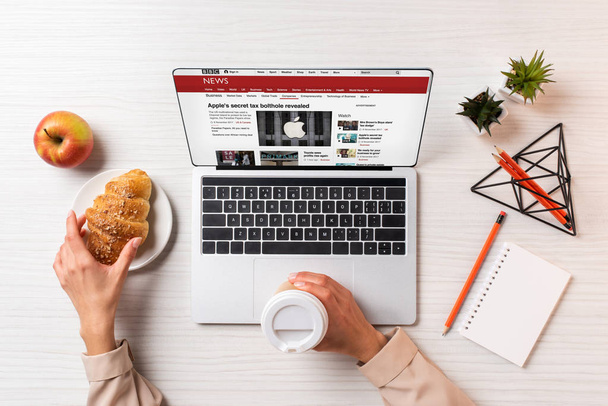 cropped shot of businesswoman holding disposable coffee cup and croissant while using laptop with bbc news website - Foto, imagen