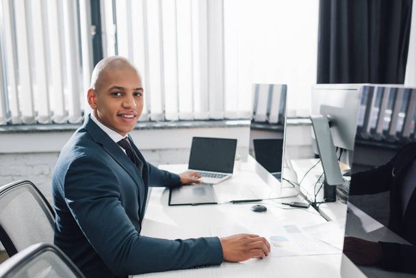 knappe jonge Afro-Amerikaanse zakenman zit op de werkplek en glimlachend in de camera in office - Foto, afbeelding