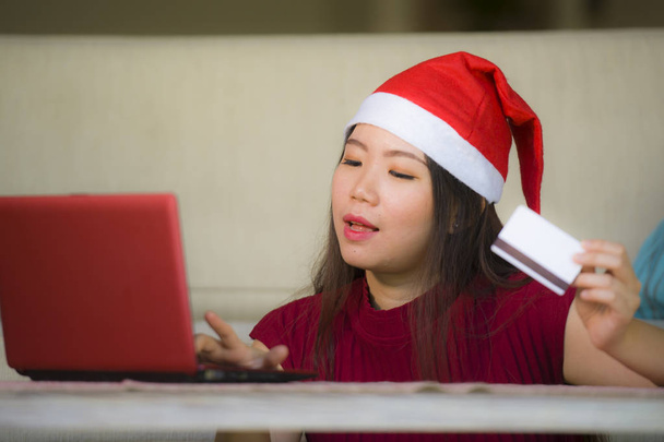young beautiful and happy Asian Korean woman in Christmas Santa Claus hat holding credit card using laptop computer for internet online shopping smiling excited at home couch living room - Photo, Image