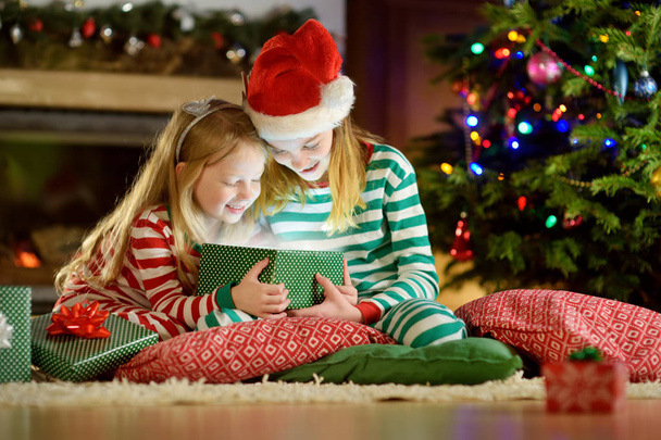 Happy little sisters wearing Christmas pajamas opening gift boxes by a fireplace in a cozy dark living room on Christmas eve. Celebrating Xmas at home. - Фото, зображення