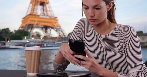 Mujer caucásica con mensajes de café en el teléfono inteligente sentado junto a la Torre Eiffel
 - Foto, Imagen