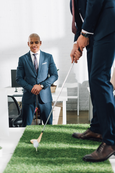 обрізаний знімок бізнесмена, який грає в гольф і посміхається афроамериканський колега, що стоїть позаду в офісі
 - Фото, зображення