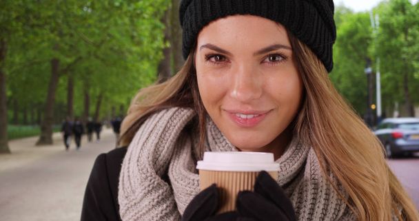 Усміхнена жінка в затишному капелюсі і шарфі тримає паперову чашку кави
 - Фото, зображення