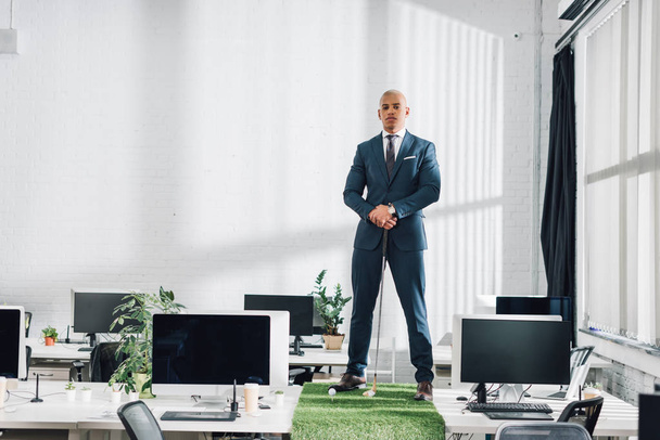 confident young african american businessman standing on table and looking at camera while playing golf in office  - Foto, Imagem