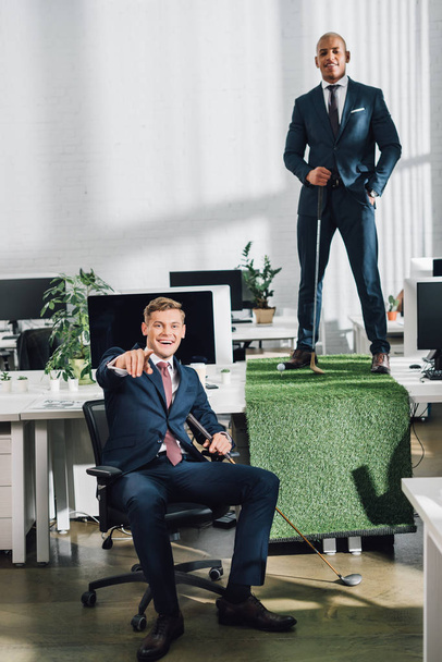 happy young multiethnic businessmen smiling at camera while playing golf in office - 写真・画像
