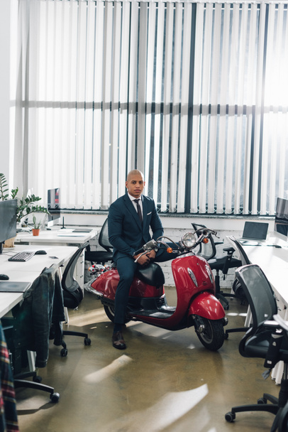handsome young african american businessman sitting on red scooter and looking at camera in office - Photo, Image