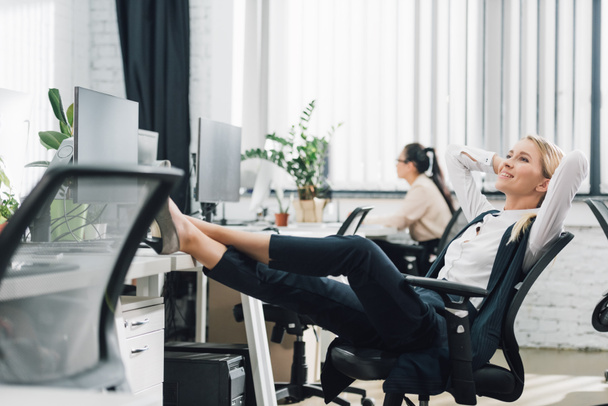 smiling young businesswoman resting with legs on desk in office - Фото, зображення