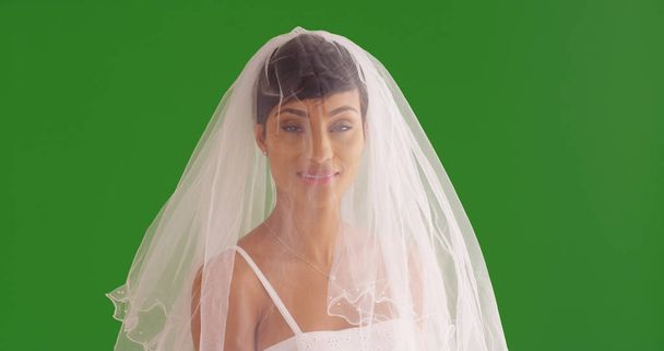 elegante Braut mit schleierbedecktem Gesicht, die auf grünem Bildschirm in die Kamera lächelt - Foto, Bild