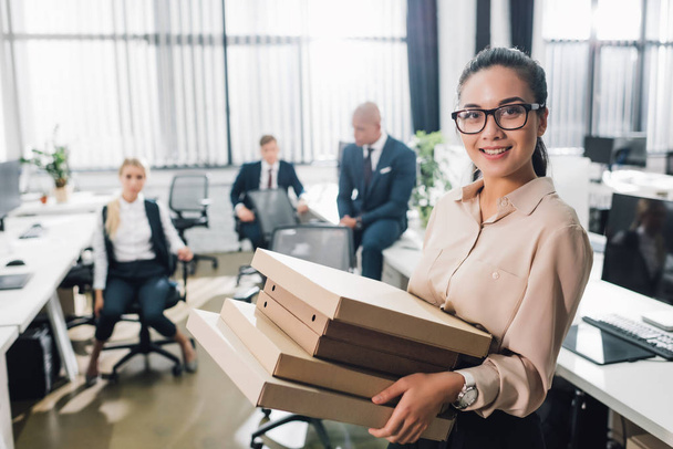 young businesswoman holding pizza boxes and smiling at camera while colleagues sitting behind in office   - Photo, Image