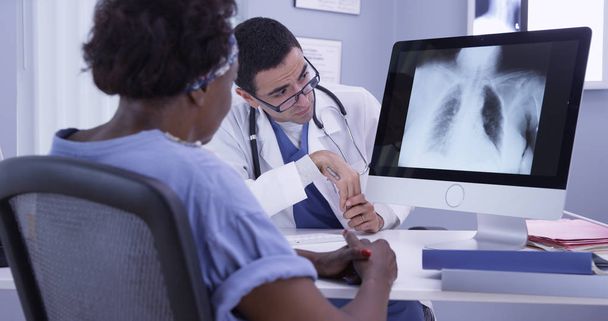 Старшая черная женщина слушает молодого латиноамериканского доктора медицины, когда он рассматривает ее рентген
 - Фото, изображение