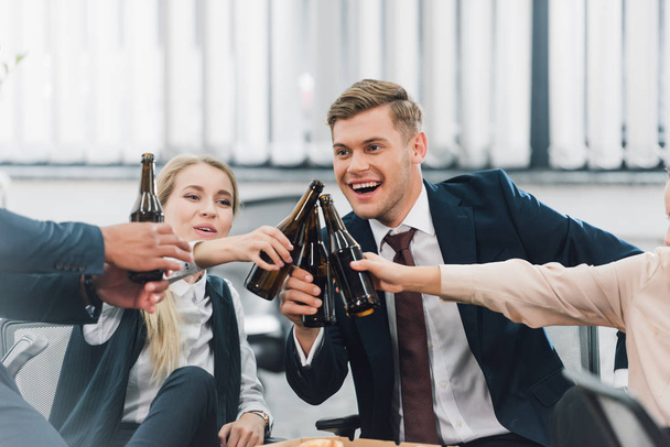 recortado tiro de feliz jóvenes compañeros de trabajo tintineo botellas de cerveza en la oficina
 - Foto, Imagen