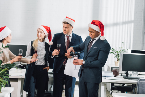 smiling young multiethnic coworkers in santa hats celebrating christmas in office   - Foto, afbeelding