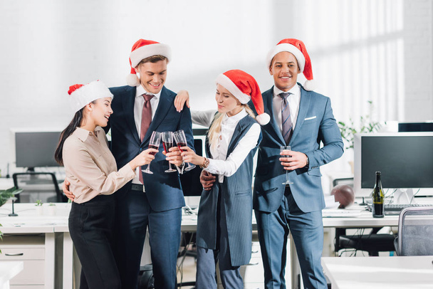 smiling young business colleagues in santa hats drinking wine and celebrating christmas in office - Photo, Image