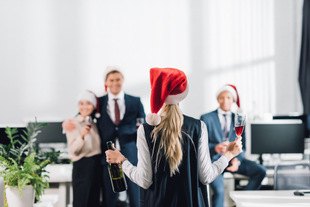 vue arrière de la femme d'affaires dans le chapeau santa tenant verre et bouteille de vin tout en célébrant Noël avec des collègues
 - Photo, image