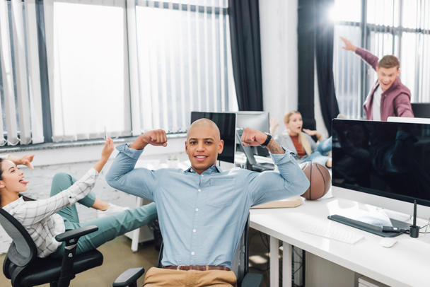 feliz joven afroamericano hombre mostrando bíceps y sonriendo a la cámara mientras sus colegas se divierten detrás en la oficina
 - Foto, imagen