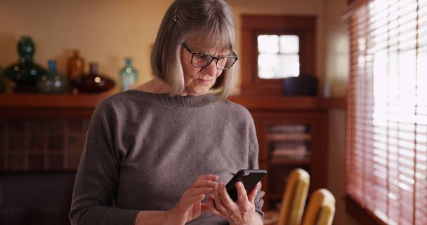 Serious senior woman using phone reading tragic news standing in living room - Photo, Image