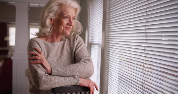 Hymyilevä vanha valkoihoinen nainen istuu ikkunan ääressä ja katselee ulos ilolla.
 - Valokuva, kuva