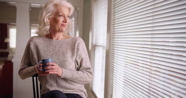 Ev içinde oturan ve pencereden dışarı bakarak kahve fincanı ile yaşlı beyaz Bayan - Fotoğraf, Görsel