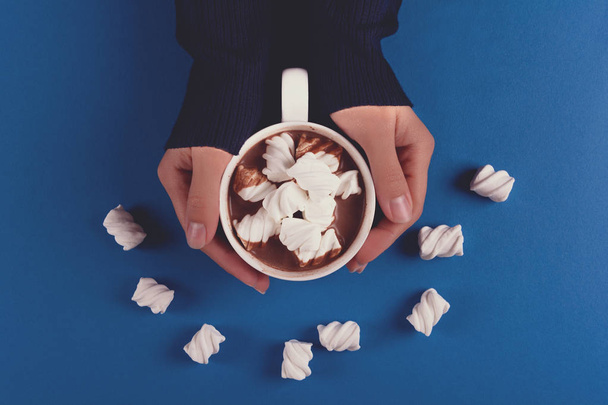 Weibliche Hände halten heiße Schokolade mit Marshmallow-Bonbons auf blauem Papierhintergrund. Ansicht von oben. Kopierraum. gemildert - Foto, Bild