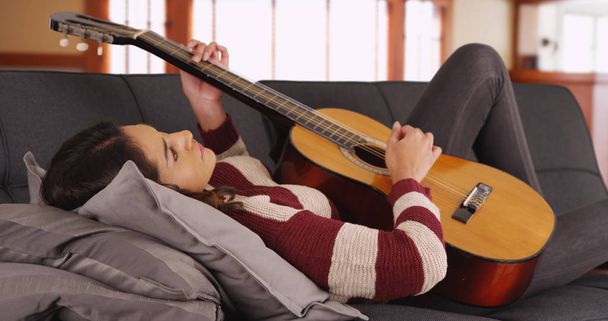 Portrait of attractive Latina female lying on sofa playing guitar and smiling - Foto, Imagen