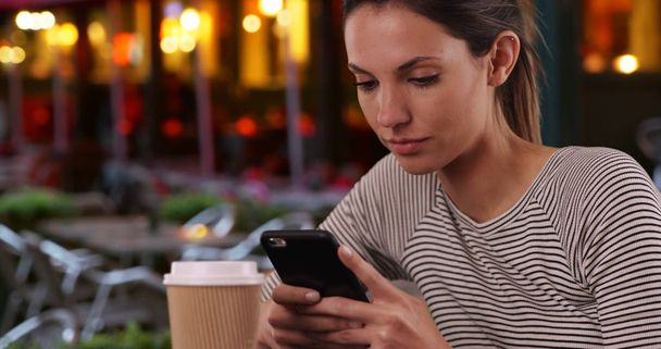 Joven dama caucásica con taza de café usando teléfono sentado al aire libre en el restaurante
 - Foto, imagen