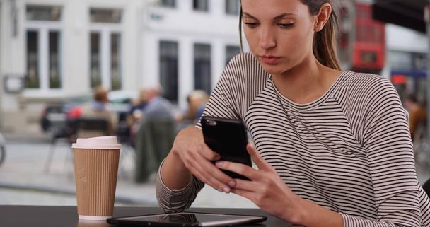Young lady sitting at cafe table texting with phone sitting at table in city - Photo, Image
