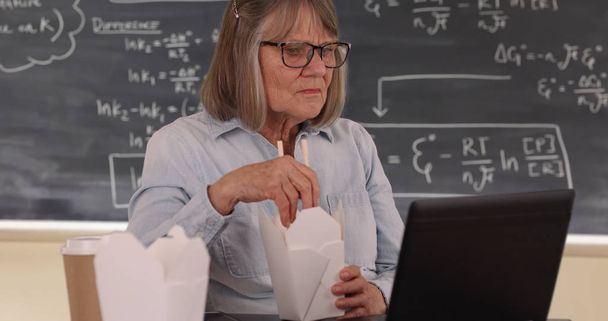 Ηλικιωμένη δασκάλα το γεύμα και χρήση φορητού υπολογιστή στην τάξη ρύθμιση - Φωτογραφία, εικόνα