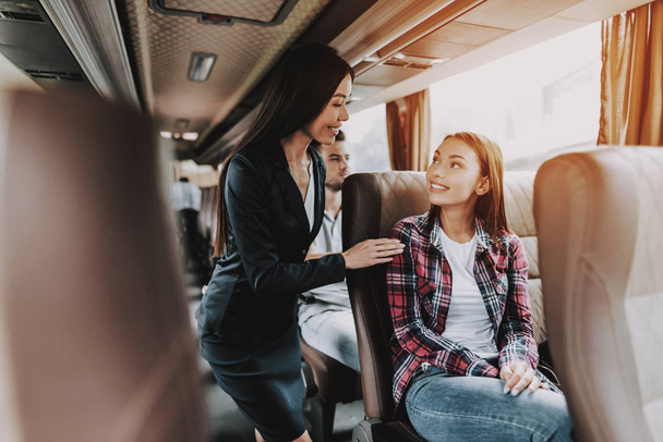 Female Tour Service Employee at Work on Tour Bus. Young Smiling Business Woman Standing between Passenger Seats and Talking to Woman. Traveling, Tourism and People Concept. People on Trip - 写真・画像