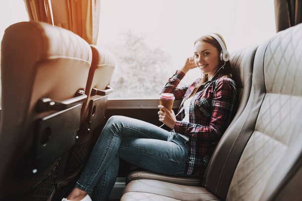 Smiling Woman Listening Music in Tourist Bus. Young Beautiful Woman Relaxing on Passenger Seat in Headphones and Drinking Coffee. Traveling, Tourism and People Concept. Happy Travelers on Trip - 写真・画像