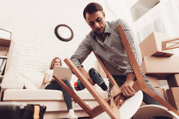 Assemble Furniture. Man Repairing Chair on Floor. Man Collects Chair. Furniture Assembler with Drill and Toolbox. Woman on Sofa with Laptop. Bright Interior. Craftsman with Tools Repairs Chair. - Photo, image