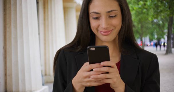 Smiling Latina businesswoman texts on smartphone outdoors - Photo, Image