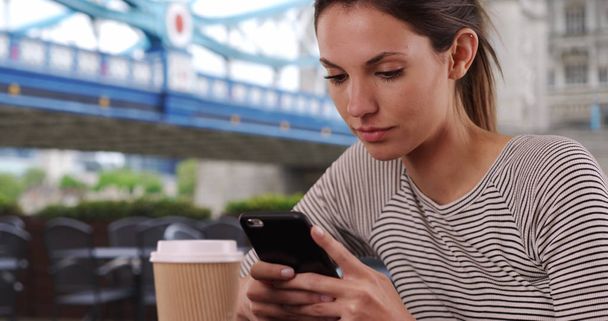 Jovem senhora caucasiana com mensagens xícara de café no telefone sentado por Tower Bridge
 - Foto, Imagem