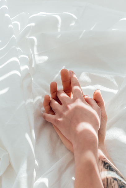 partial view of loving couple holding hands while lying on white bed sheet - Valokuva, kuva