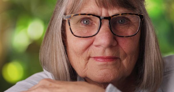 Nahaufnahme einer entspannten Seniorin, die in üppiger Outdoor-Umgebung in die Kamera blickt - Foto, Bild