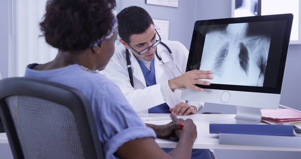 Senior negro mujer escuchando a joven latino MD como él revisa su rayos X
 - Foto, imagen