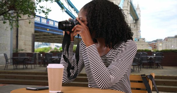 Musta nainen istuu ulkona kahvilassa lähellä Tower Bridgeä ottamassa kuvia kameralla
 - Valokuva, kuva