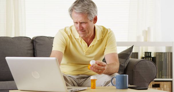 Keski-ikäinen mies pitelee lääkepulloa ja lukee läppäriään
  - Valokuva, kuva
