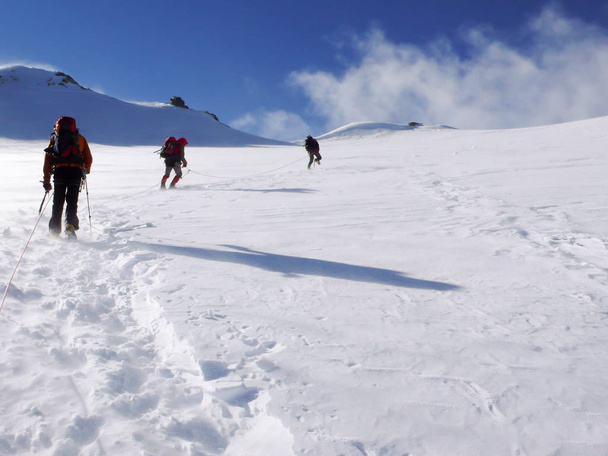 three mountain climbers cross a high alpine glacier in windy stormy weather on their way to a mountain peak far ahead - Fotoğraf, Görsel