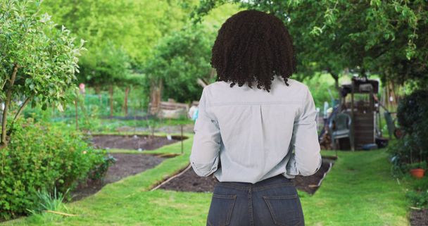 Задний вид на молодую чернокожую женщину, оглядывающуюся по сторонам в саду
 - Фото, изображение