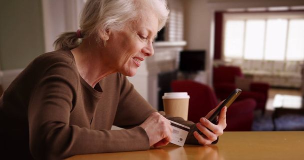 現代の年配の女性の家の中に座っている彼女の携帯電話を介してオンライン ショッピング - 写真・画像