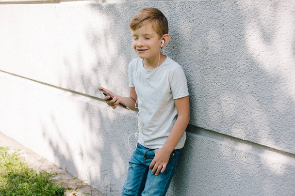 Fiatal fiú viselésével fülhallgatók, keres el. 7 éves fiú a smartphone. Gyermek szőke fiatal fiú szabadban portréja. zenehallgatás fejhallgató - Fotó, kép