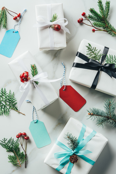 κάτοψη του λευκά χριστουγεννιάτικα δώρα με κλαδιά έλατου, διακοσμητικά μούρα και ετικέτες στο μαρμάρινο τραπέζι  - Φωτογραφία, εικόνα