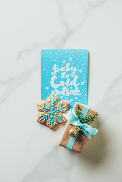 top view of christmas present, snowflake cookie and greeting card with "baby its cold outside" lettering on marble background - Photo, Image