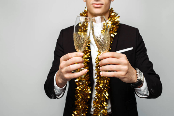 attractive man in a business suit, and with a tinsel around his neck, a man holding a glass of champagne, on a gray background. The concept of the new year. - Zdjęcie, obraz