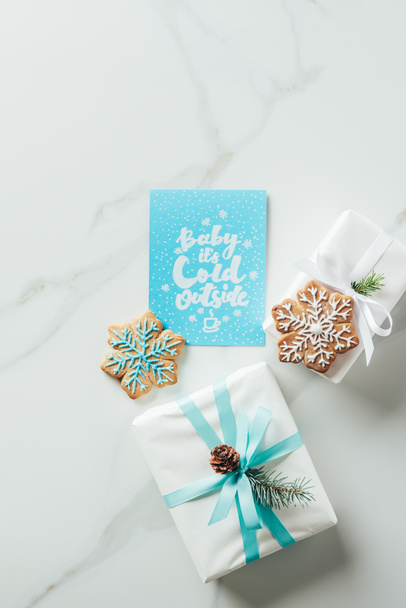 top view of white christmas gifts, snowflake cookies and greeting card with "baby its cold outside" lettering on marble table - Zdjęcie, obraz