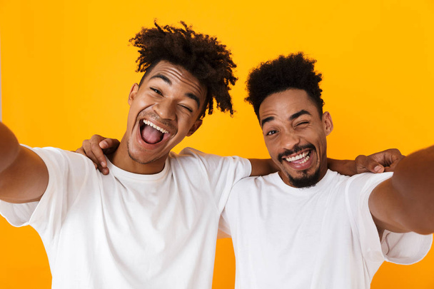 Two joyful male african friends in t-shirts standing isolated over yellow background, taking a selfie - Fotografie, Obrázek